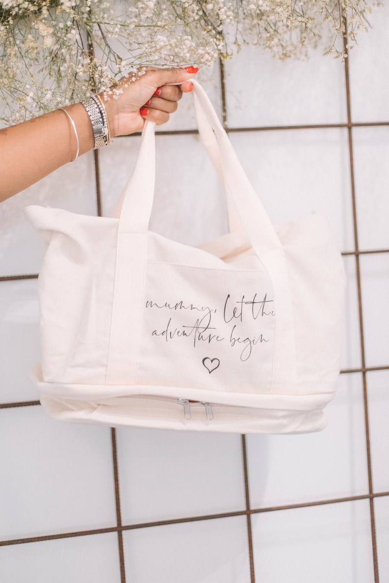Op de afbeelding: Een witte canvas tote bag met een ritssluiting. De tas heeft een zwarte inscriptie met de tekst "mummy, let the adventure begin" met een klein hartje onder de tekst.