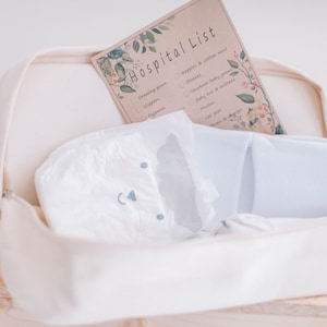 Op de afbeelding: Een witte canvas tas met een ritssluiting, die een luier, een paar witte pantoffels en een geprinte checklist met de titel "Hospital List" bevat.