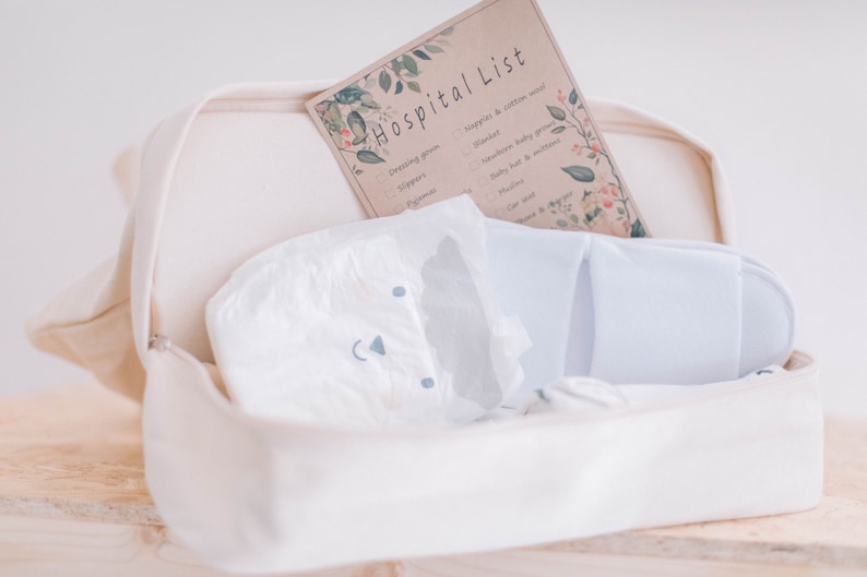 Op de afbeelding: Een witte canvas tas met een ritssluiting, die een luier, een paar witte pantoffels en een geprinte checklist met de titel "Hospital List" bevat.