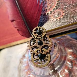 Peut inclure: Un élément décoratif sur le thème steampunk avec une plume bordeaux et des engrenages en bronze. Les engrenages sont superposés et fixés à un objet en verre transparent. Un journal avec des engrenages en relief est visible en arrière-plan.