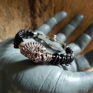 Peut inclure: Un bracelet en cuir noir avec une pièce maîtresse décorative argentée représentant une coiffe amérindienne ornée. Le bracelet a une bande en cuir tressé et un fermoir argenté.