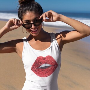 May include: A woman wearing a white tank top with a red sequined lips design and black bikini bottoms. She is standing on a sandy beach with the ocean in the background.