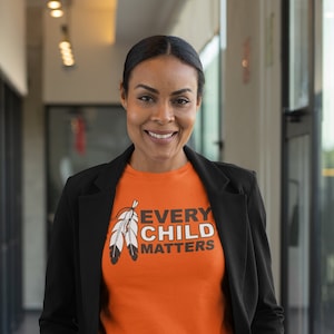 May include: A person wearing a black blazer over an orange t-shirt with the text "Every Child Matters" and a white feather design. The person is standing in a hallway.