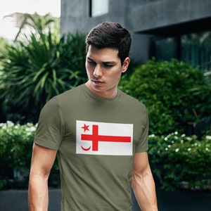 May include: A man wearing a green t-shirt with a white and red flag design. The flag has a red cross, a red star, and a crescent moon.