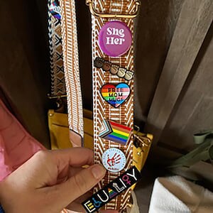 May include: A brown and white patterned bag strap with several pins attached. The pins include a rainbow heart with the text "Free Mom Hugs", a rainbow stripe with a brown shape, a white circle with a red handprint, a pink circle with the text "She Her", and a black and white rectangle with the text "Equality+".