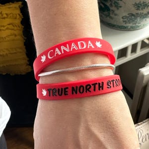 May include: Two red silicone bracelets with white lettering. The top bracelet says "CANADA" with a maple leaf on each side. The bottom bracelet says "TRUE NORTH STRONG" with a maple leaf on the left side.