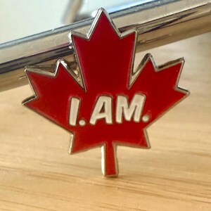 May include: A red maple leaf pin with a white border and the words "I AM." in white letters.