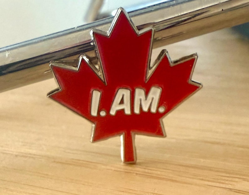 May include: A red maple leaf pin with a white border and the words "I AM." in white letters.