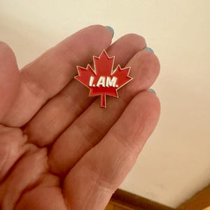 May include: A red maple leaf pin with a white outline and the words "I AM." in white letters.