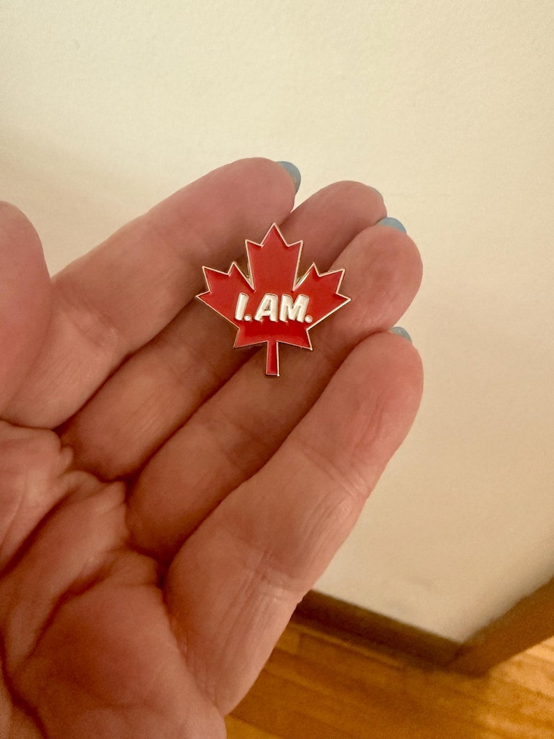 May include: A red maple leaf pin with a white outline and the words "I AM." in white letters.