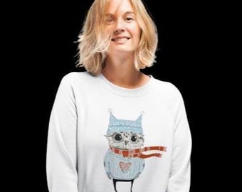 May include: A white long-sleeve sweatshirt featuring a graphic illustration of an owl wearing a light blue knit hat and a matching sweater with a red heart. The owl is also wearing a long, flowing red and white striped scarf around its neck.
