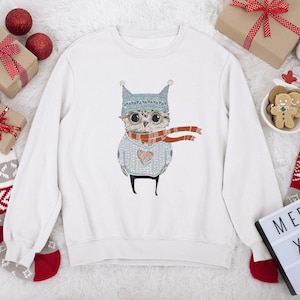 May include: White sweatshirt with a Christmas owl design. The owl is wearing a blue hat and a red scarf. The sweatshirt is surrounded by Christmas decorations, including stockings, ornaments, and a sign that says "Merry Xmas".