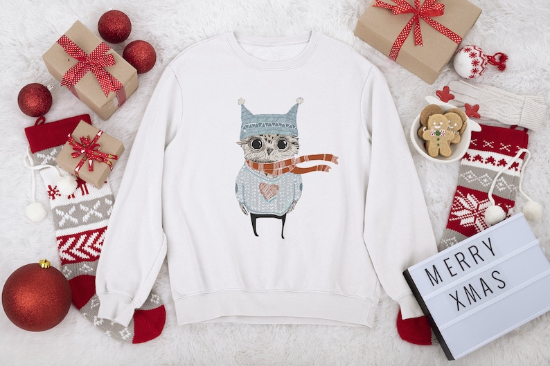May include: White sweatshirt with a Christmas owl design. The owl is wearing a blue hat and a red scarf. The sweatshirt is surrounded by Christmas decorations, including stockings, ornaments, and a sign that says "Merry Xmas".