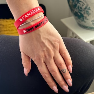 May include: Two red silicone bracelets on a person's wrist. The top bracelet says "CANADA" with a maple leaf on each side. The bottom bracelet says "TRUE NORTH STRONG" with a maple leaf on the left side.