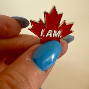 May include: A red maple leaf pin with the words "I AM" in white lettering.