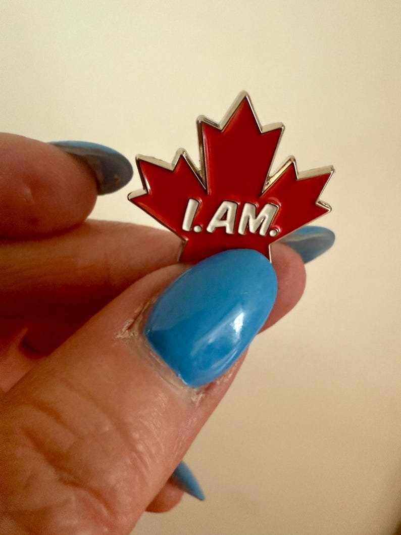 May include: A red maple leaf pin with the words "I AM" in white lettering.