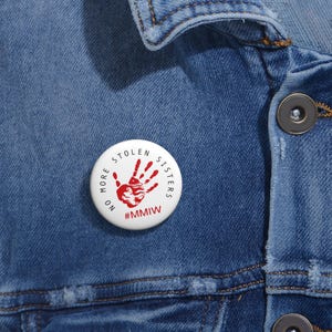 May include: A white button badge with red text and a handprint design. The text around the edge reads "NO MORE STOLEN SISTERS", with the hashtag "#MMIW" at the bottom. The badge is on a denim jacket.