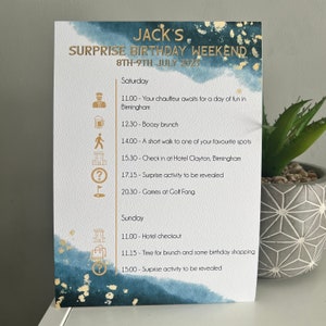 May include: A blue and gold illustrated card with the text "Jack's Surprise Birthday Weekend 8th-9th July 2023". The card lists a weekend itinerary with activities including a chauffeur, boozy brunch, a walk to a favourite spot, checking into a hotel, a surprise activity, and games at Golf Fang.
