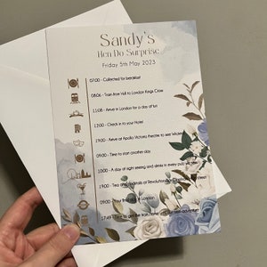 May include: A light blue and white floral design card with a gold border. The card lists a schedule of events for a hen do surprise in London, England. The schedule includes times and activities such as collecting for breakfast, train travel, arriving in London, checking into a hotel, seeing a show, and having drinks at a pub.