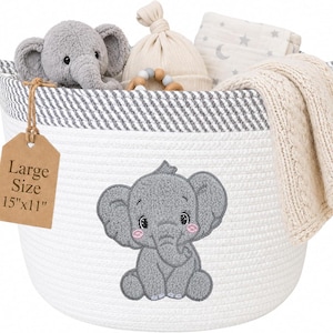 Op de afbeelding: Een witte touw opbergmand met een grijze gestreepte rand en een grijze olifant borduursel. Een bruin label toont Large Size 15"x11". Binnenin zitten een grijze pluche olifant, een beige knoopmuts, een houten kralen bijtring en een crèmekleurige deken.