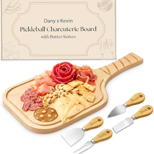 May include: A light wood charcuterie board shaped like a pickleball paddle, featuring a carved pickleball design, topped with salami, prosciutto, cheese, and crackers. Four matching wood-handled knives are labeled Smash, Dink, Serve, and Rally.