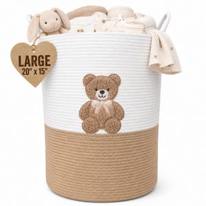 Op de afbeelding: Een grote opbergmand van wit en beige touw met een bruine teddybeer-applicatie aan de voorzijde. Een hartvormig label aan de zijkant toont de tekst LARGE 20" x 15". De mand is gevuld met diverse babyartikelen en een knuffeldier.