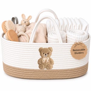 May include: An oval woven rope storage basket with white and tan sections, featuring a brown teddy bear graphic on the front and a tag reading Removable Dividers. The basket contains baby items including wooden brushes, a rattle, and folded cloths.