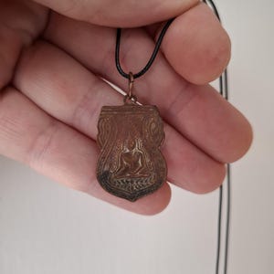 May include: A copper pendant necklace with a detailed design of a seated Buddha figure. The pendant is attached to a black cord.