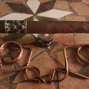 May include: Two copper cigar rests, one is a simple ring, the other is a more elaborate design with a loop and a curved arm. A brown cigar with a black and white label is resting on a brown tiled surface.