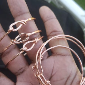 May include: Several copper-colored bangles and bracelets. Some are simple, round bands, while others feature a knot design with an oval-shaped loop. The bracelets are displayed on a hand, showcasing the jewelry's color and design.