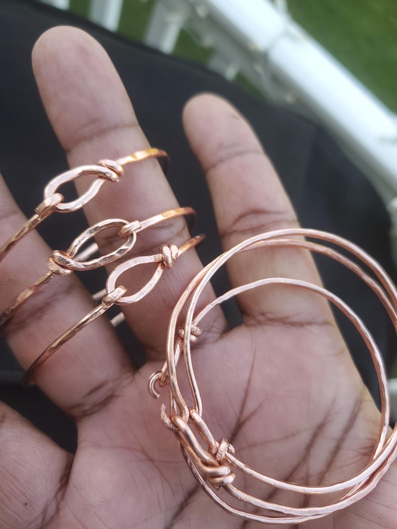 May include: Several copper-colored bangles and bracelets. Some are simple, round bands, while others feature a knot design with an oval-shaped loop. The bracelets are displayed on a hand, showcasing the jewelry's color and design.