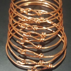 May include: A stack of copper-colored bangles. Each bangle is a simple, round band with a knot-like closure. The metal has a warm, reddish-brown hue, and the design is minimalist. The bangles are stacked on a dark surface.