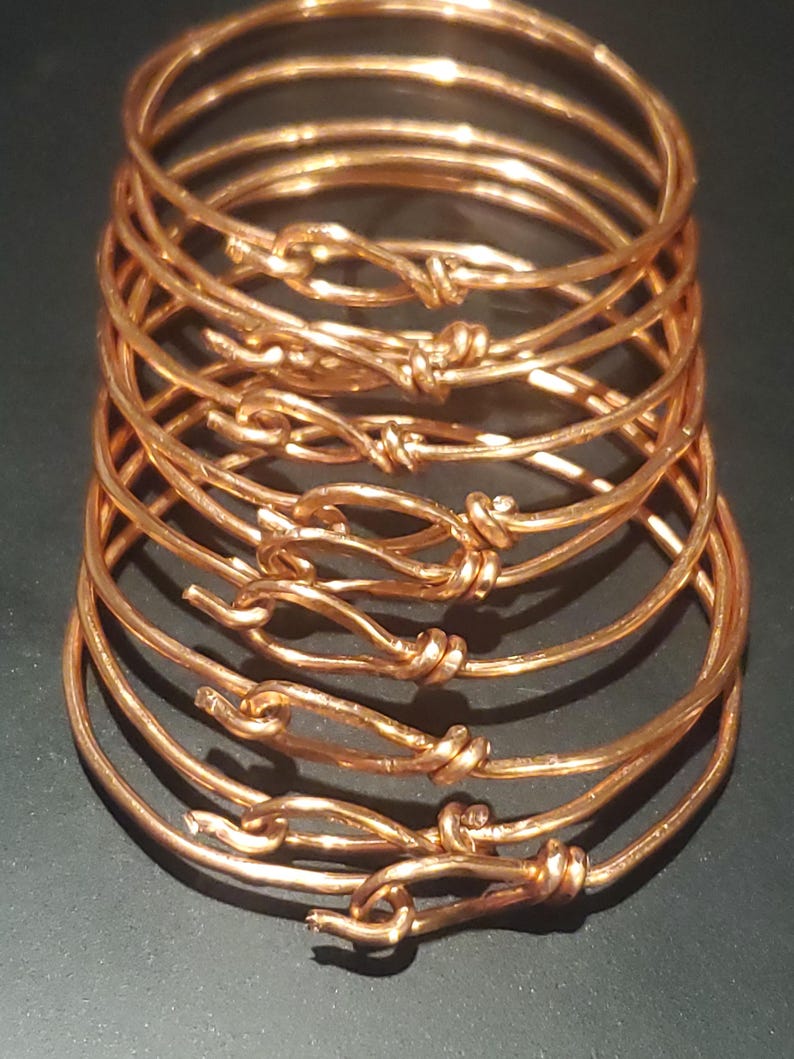 May include: A stack of copper-colored bangles. Each bangle is a simple, round band with a knot-like closure. The metal has a warm, reddish-brown hue, and the design is minimalist. The bangles are stacked on a dark surface.
