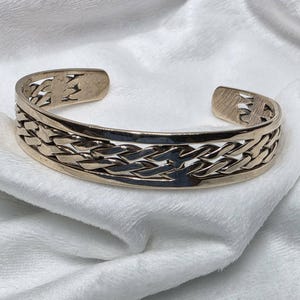May include: A gold-toned cuff bracelet with a woven design. The bracelet has an open design and is resting on a white, textured surface. The woven pattern is the focal point of the bracelet, adding a touch of elegance.