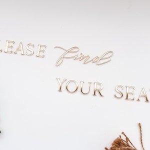 May include: Gold-colored metal lettering spelling out "PLEASE Find YOUR SEAT" on a white background. The word "Find" is in a cursive font. Green leaves and brown twine are in the corners.