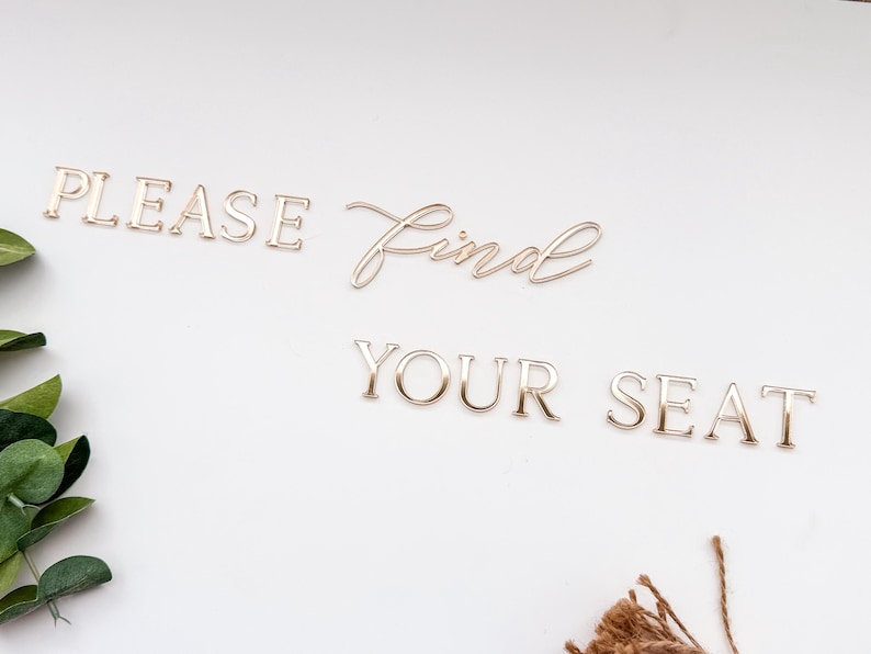 May include: Gold-colored metal lettering spelling out "PLEASE Find YOUR SEAT" on a white background. The word "Find" is in a cursive font. Green leaves and brown twine are in the corners.