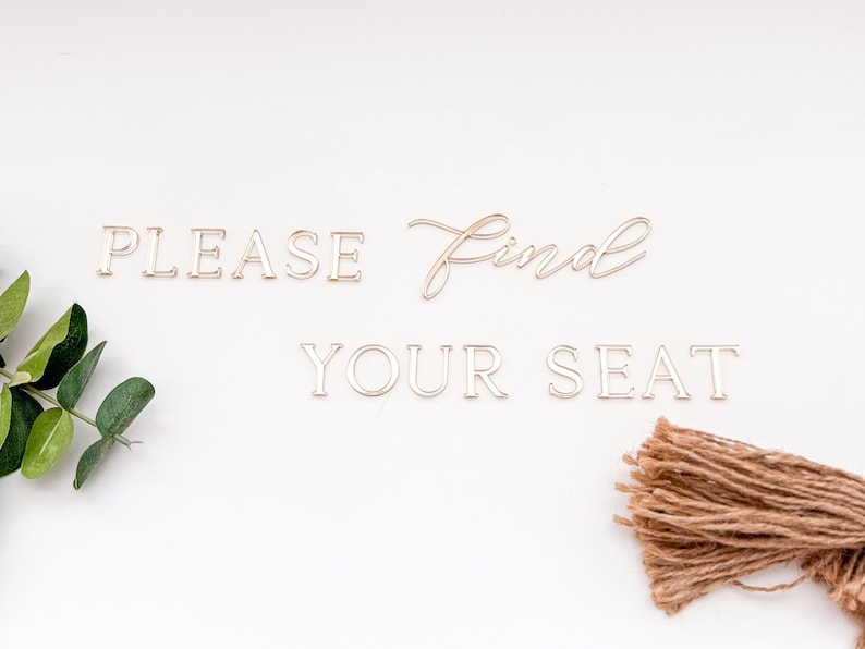 May include: Gold-colored metal letters spell out "PLEASE FIND YOUR SEAT" on a white background. The word "find" is in cursive script. Green leaves and a bundle of brown twine are in the corners.