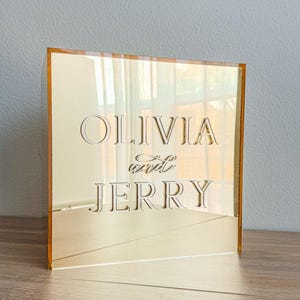 May include: A gold-toned acrylic sign with the names "OLIVIA" and "JERRY" engraved in a classic font. The sign is square and stands upright, reflecting the surrounding environment. The edges have a gold border.