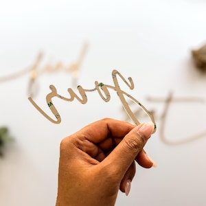 May include: A gold-colored, cursive word "sure" is held in a hand. The word has a double-sided adhesive on the back. Other gold-colored words are blurred in the background. The background is white.