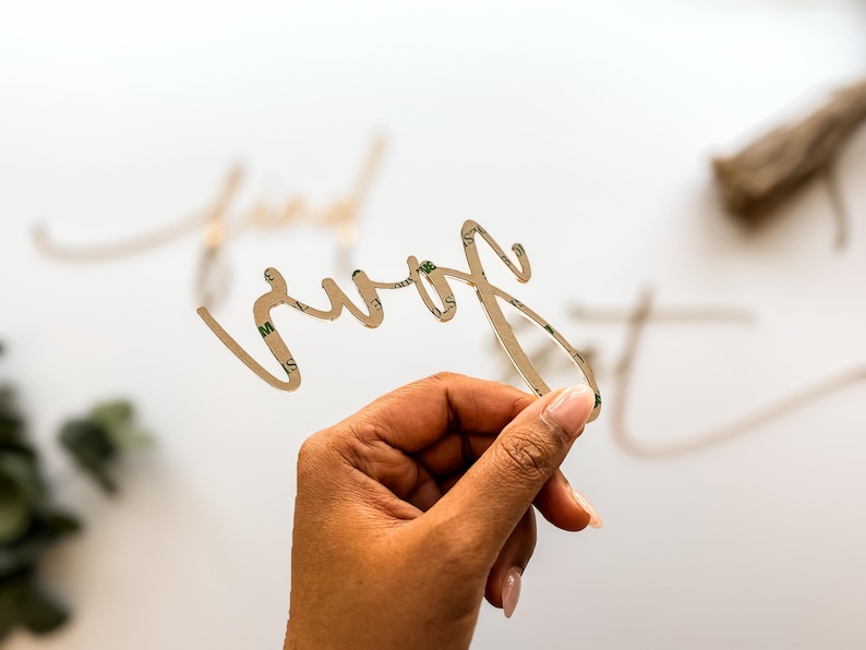 May include: A gold-colored, cursive word "sure" is held in a hand. The word has a double-sided adhesive on the back. Other gold-colored words are blurred in the background. The background is white.