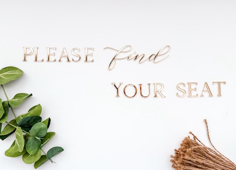 May include: Gold-colored metal letters spell out "PLEASE Find YOUR SEAT" on a white background. Green leafy sprigs and a bundle of twine are arranged around the text, creating a decorative wedding or event sign.