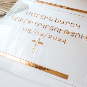 May include: Clear acrylic box with gold trim and engraved text. The text includes a date and a cross symbol. The box is designed for a special occasion, possibly a baptism or christening, and is displayed on a white surface.