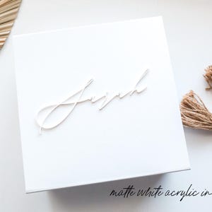 May include: A white square box with the name "Joseph" in a cursive font. The text "matte white acrylic in font #7" is at the bottom right. Decorative elements include a dried palm leaf and a beaded tassel.