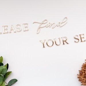 May include: Gold-colored metal lettering spelling "PLEASE FIND YOUR SEAT" on a white background. The word "find" is in a cursive font. Green leaves and a bundle of twine are in the corners.