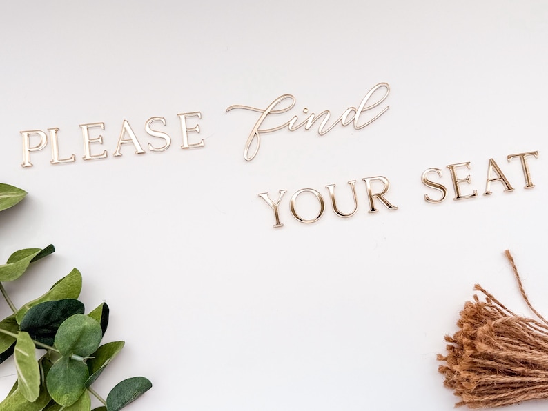 May include: Gold-colored metal lettering spelling "PLEASE FIND YOUR SEAT" on a white background. The word "find" is in a cursive font. Green leaves and a bundle of twine are in the corners.