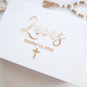 May include: A white box with gold lettering that reads "Lucas" and "October 15, 2023" with a gold cross below. A wooden rosary and a baby toy are in the background.