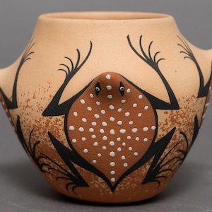 May include: A small, terracotta pottery vase with a brown and black frog design. The frog has white dots on its back and is facing forward. The vase has a rounded shape with a wide opening.