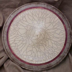 May include: A round ceramic plate with a white glaze and a crackle finish. The plate has a dark brown rim with a thin red line around the edge.