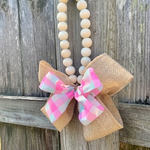 May include: A wooden bead garland with a pink and white checkered bow and burlap ribbon. The garland is hanging on a wooden fence.