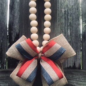 May include: A wooden bead garland with a red, white, and blue burlap bow. The garland is hanging on a wooden fence.